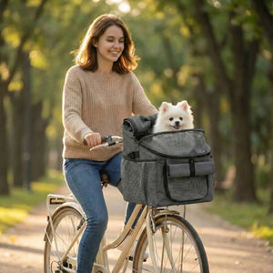 Une femme souriante fait du vélo dans un parc ensoleillé, transportant un petit chien blanc pelucheux dans un panier de velo pour petit chien gris fixé au guidon.